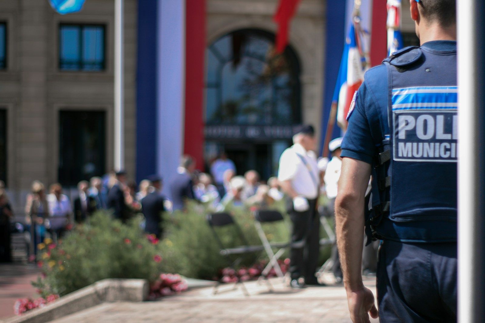 Policier municipal au premier plan. Plusieurs personnes au second plan, devant un bâtiment avec le drapeau français.