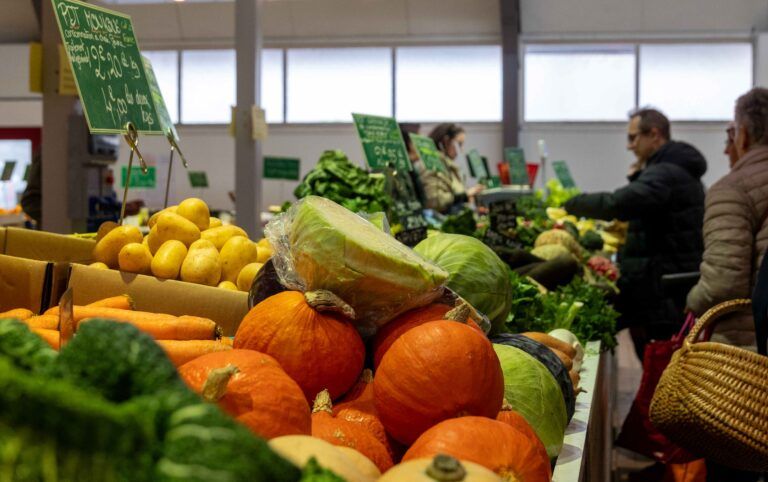 Intérieur du Marché des Saveurs avec un zoom sur un étal de fruits et légumes et des clients en arrière plan
